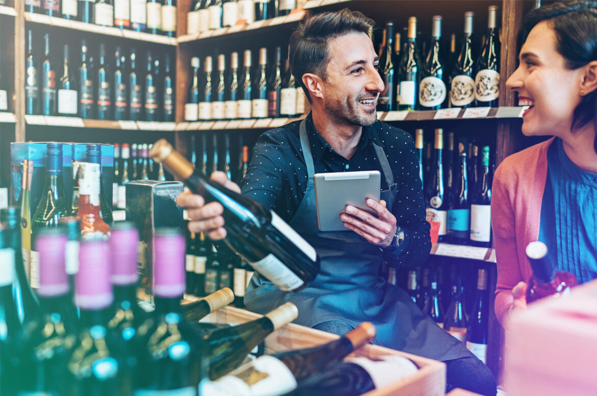 A liquor store business owner is holding a wine bottle while discussing with a client.